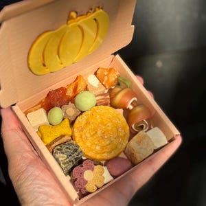 May include: A wooden box filled with assorted dog treats. The box has a pumpkin-shaped embellishment on the lid. The treats are various shapes and colors, including yellow, green, and brown.