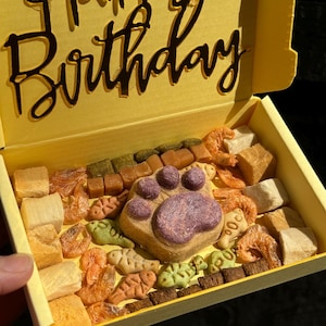 May include: A yellow box with the words "Happy Birthday" on the lid. The box is filled with a variety of dog treats, including a large paw-shaped treat in the center.