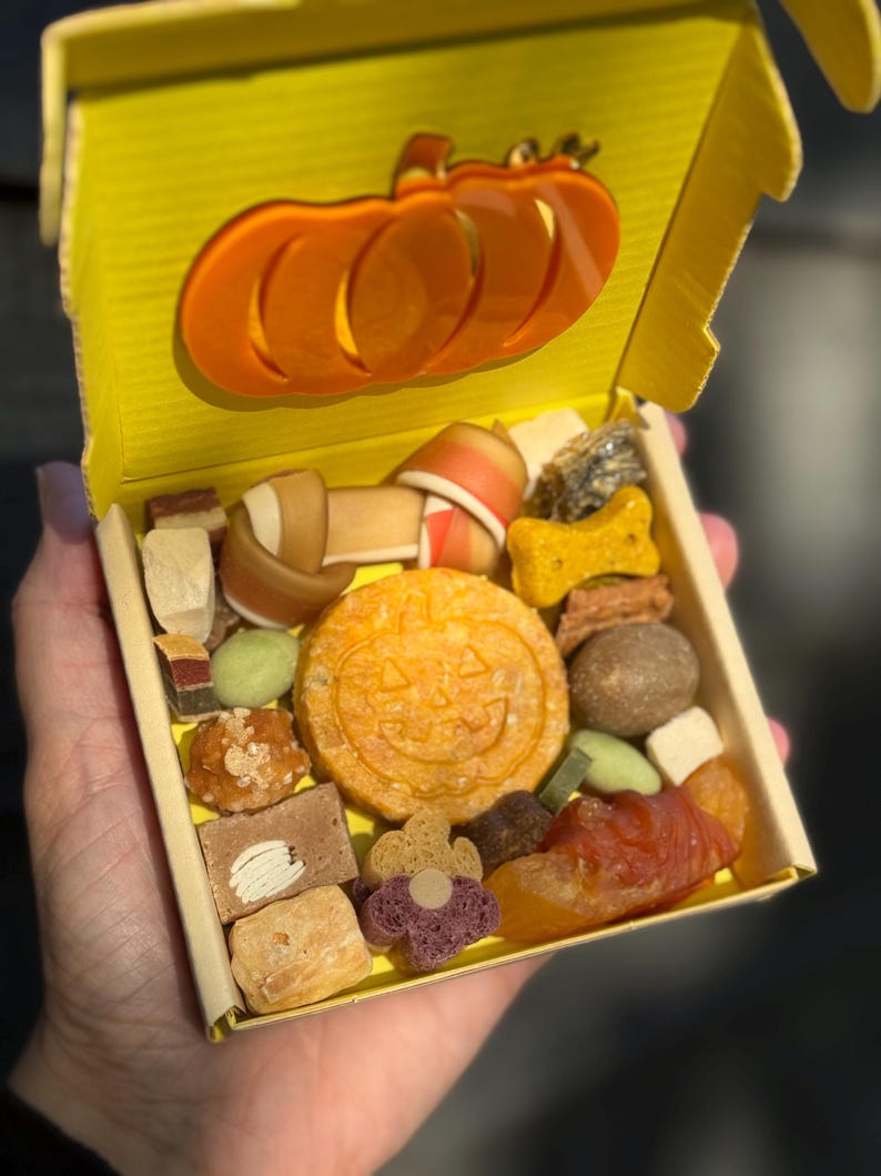 May include: A yellow box filled with various dog treats, including a pumpkin-shaped biscuit and bone-shaped chews. The box has an orange pumpkin cutout on the lid. The treats are in different shapes and colors.