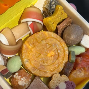May include: A wooden box filled with various dog treats. The treats include a bone-shaped biscuit, a round biscuit with a jack-o'-lantern face, and other colorful snacks. The box is yellow inside and has an orange pumpkin shape on the side.