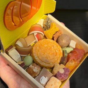 May include: A yellow box filled with various dog treats, including a pumpkin-shaped cookie and bone-shaped biscuits. An orange pumpkin-shaped charm is attached to the box. The treats are in different colors and textures.