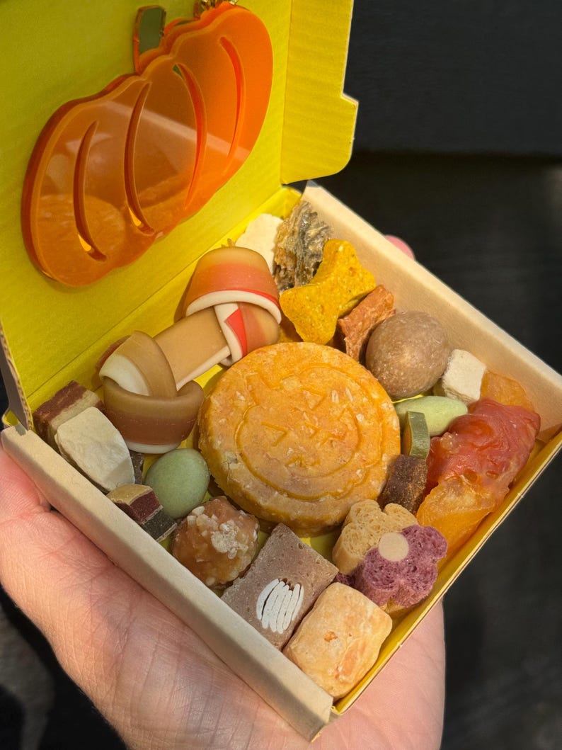 May include: A yellow box filled with various dog treats, including a pumpkin-shaped cookie and bone-shaped biscuits. An orange pumpkin-shaped charm is attached to the box. The treats are in different colors and textures.