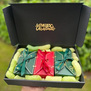 May include: A black gift box with a gold "Merry Christmas" embellishment. Inside, three wrapped gift boxes in red and green are nestled in green packing material. Each gift is tied with a ribbon that says "For You".