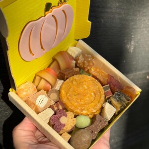 May include: A yellow box with a pink pumpkin cutout on the lid, filled with a variety of dog treats. The treats include bone-shaped chews, round biscuits, and other colorful snacks. The box is held in a person's hand.