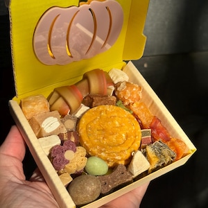 May include: A yellow box with a pink pumpkin cutout on the lid, filled with various dog treats. The treats are in different shapes and colors, including a large round orange treat and a bone-shaped treat.