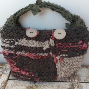 Puede incluir: Un bolso de mano tejido en color marrón, beige y rosa con un asa de tela verde y dos botones de madera.