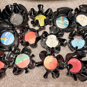 Puede incluir: Una colección de cuencos negros de discos de vinilo fundidos, cada uno con una etiqueta de disco colorida. Los cuencos tienen formas orgánicas únicas, mostrando las etiquetas originales de los discos con varios diseños y colores. Un artículo de decoración del hogar creativo y reciclado.