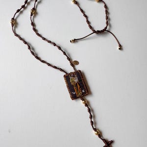 Puede incluir: Un collar de cordón anudado marrón con cuentas doradas y un pequeño colgante rectangular con una imagen religiosa. El colgante está unido a una cruz dorada.