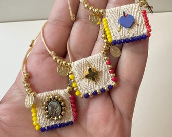 May include: Three bracelets with colorful beads and charms. Each bracelet has a small fabric square with a different design: a heart, a star, and a picture of a saint. The bracelets are tied with a string and have gold beads.