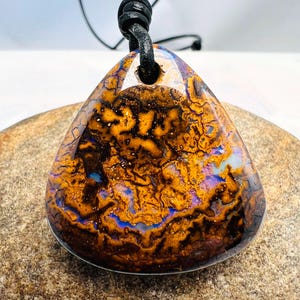 May include: A triangular boulder opal pendant featuring a polished surface with intricate patterns of deep brown, vibrant orange, and iridescent flashes of purple and blue. The stone is suspended from a black leather cord with a knotted loop.