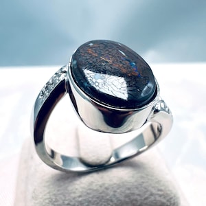 May include: A silver ring with a large oval-shaped opal gemstone set in a bezel setting. The opal is a dark blue-grey color with a pattern of white and brown veins. The ring band is decorated with small diamonds.
