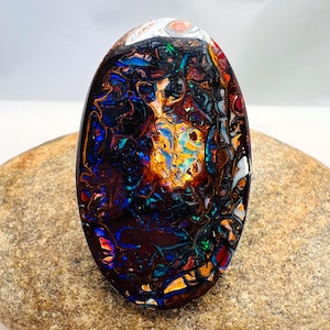 Peut inclure: Une pierre précieuse opale boulder de forme ovale présentant une matrice en pierre de fer brun foncé avec des éclats vibrants de bleu, vert, orange et jaune irisés sur sa surface polie et réfléchissante, révélant un motif géologique naturel.