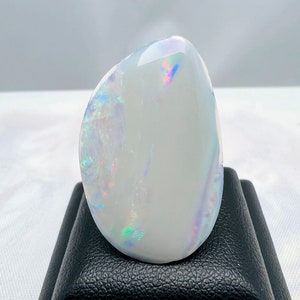 May include: A polished, oval-shaped opal gemstone. The opal has a milky white base color with iridescent flashes of blue, green, and pink. The gemstone is displayed on a black stand, showcasing its smooth, reflective surface.