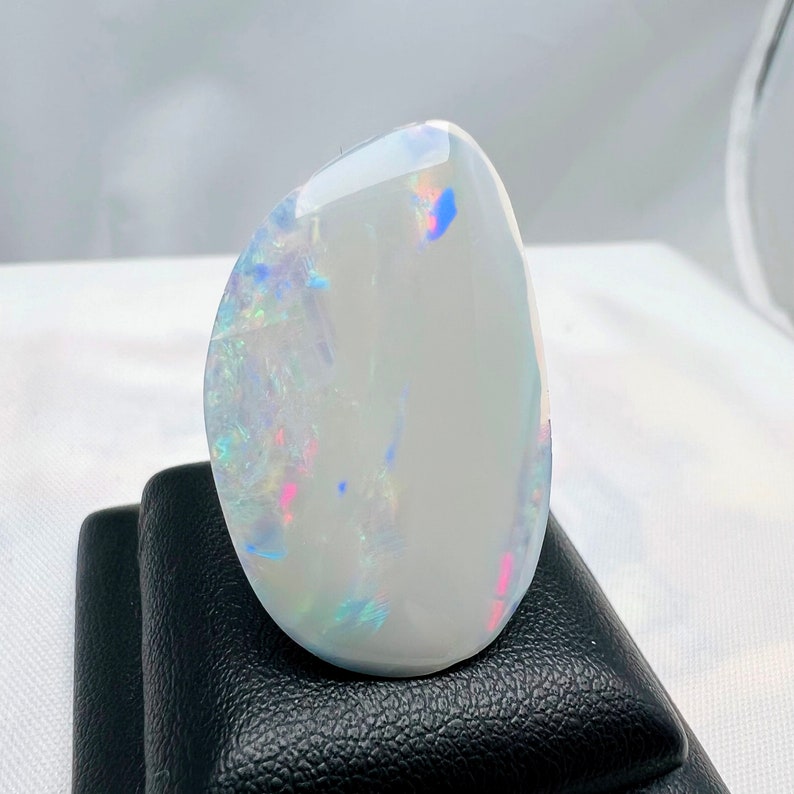 May include: A polished, oval-shaped gemstone with a milky white base and iridescent flashes of color. The stone displays a play of light with hues of pink, blue, and green. It is set on a black display stand.