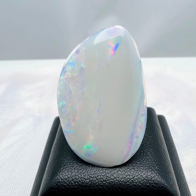 May include: A polished, oval-shaped opal gemstone. The opal has a milky white base color with iridescent flashes of blue, green, and pink. The gemstone is displayed on a black stand, showcasing its smooth, reflective surface.