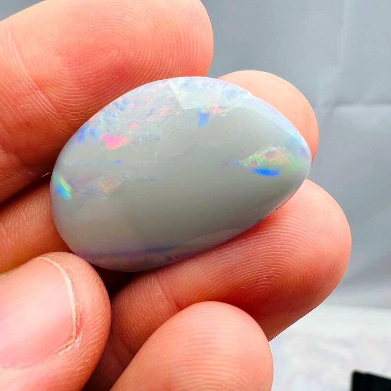 May include: A polished, oval-shaped opal gemstone. The opal has a milky white base color with iridescent flashes of pink, blue, and green across its surface. The gemstone is held in a hand, showcasing its smooth, reflective qualities.