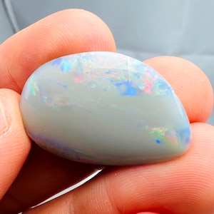 May include: A polished, oval-shaped opal gemstone. The opal has a milky white base color with iridescent flashes of blue, pink, and green across its surface. The gemstone is held between two fingers, showcasing its smooth, reflective texture.