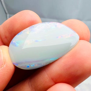 May include: A polished, oval-shaped opal gemstone. The opal has a milky white base color with iridescent flashes of blue, pink, and green. The gemstone is held between fingers, showcasing its smooth, reflective surface.