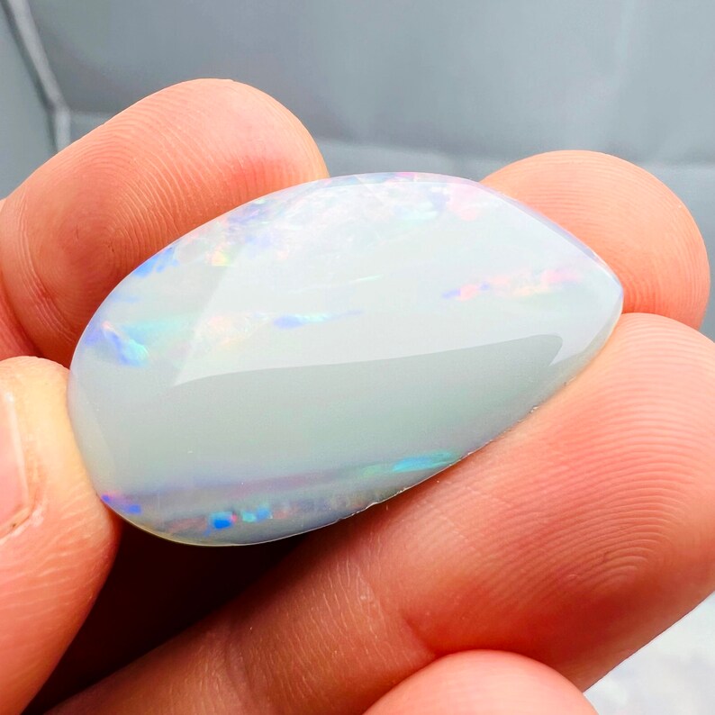 May include: A polished, oval-shaped opal gemstone. The opal has a milky white base color with iridescent flashes of blue, pink, and green. The gemstone is held between fingers, showcasing its smooth, reflective surface.