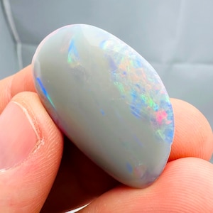 May include: A polished, oval-shaped opal gemstone. The opal has a light gray base color with iridescent flashes of blue, green, and pink across its surface. The stone is smooth and appears to be held in a hand.
