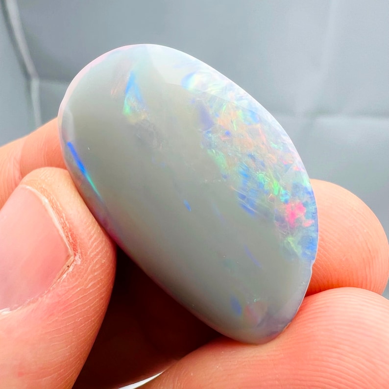 May include: A polished, oval-shaped opal gemstone. The opal has a light gray base color with iridescent flashes of blue, green, and pink across its surface. The stone is smooth and appears to be held in a hand.