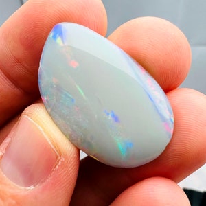 May include: A polished, teardrop-shaped opal gemstone. The opal has a milky white base color with iridescent flashes of blue, pink, and green. The stone is held between fingers, showcasing its smooth, reflective surface and natural beauty.
