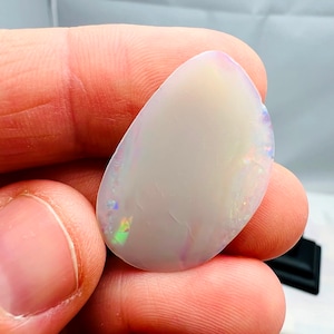 May include: A polished, teardrop-shaped opal gemstone. The opal has a milky white base color with iridescent flashes of green, blue, and purple. The stone is held between fingers, showcasing its smooth surface and natural beauty.