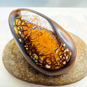 Könnte beinhalten: Ein brauner und goldener Opal mit schillernden blauen und grünen Blitzen, eingebettet in ein glattes, oval geformtes Stück polierten Gesteins.