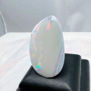 May include: A polished, oval-shaped opal with a predominantly white base color. The opal displays iridescent flashes of color, including red, blue, and green, across its surface. The gemstone is set against a black background.
