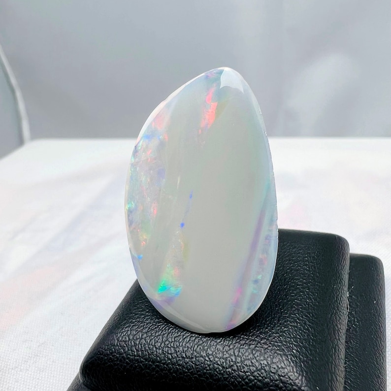 May include: A polished, oval-shaped opal with a predominantly white base color. The opal displays iridescent flashes of color, including red, blue, and green, across its surface. The gemstone is set against a black background.