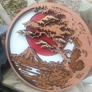 Puede incluir: Arte mural redondo de madera que representa una montaña, un árbol y un sol rojo. El diseño utiliza madera en capas para crear un efecto 3D. La obra de arte es predominantemente marrón, con un fondo blanco y rojo. El árbol y la montaña están intrincadamente cortados.
