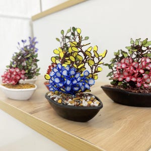 May include: Miniature bonsai trees with colorful, handcrafted floral arrangements. The bonsai feature red, blue, yellow, and purple flowers, set in small, oval-shaped pots. The pots are filled with small stones and sit on a wooden shelf.