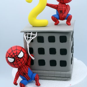 Puede incluir: Un pastel de dos pisos decorado con un n&uacute;mero dos amarillo y dos figuras de Spiderman rojas y azules. El piso inferior es un edificio gris con ventanas negras. El pastel est&aacute; sobre una superficie blanca.