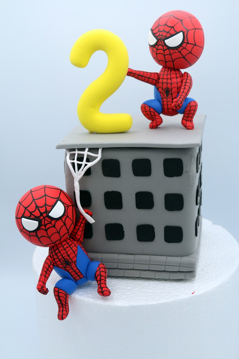 Puede incluir: Un pastel de dos pisos decorado con un n&uacute;mero dos amarillo y dos figuras de Spiderman rojas y azules. El piso inferior es un edificio gris con ventanas negras. El pastel est&aacute; sobre una superficie blanca.