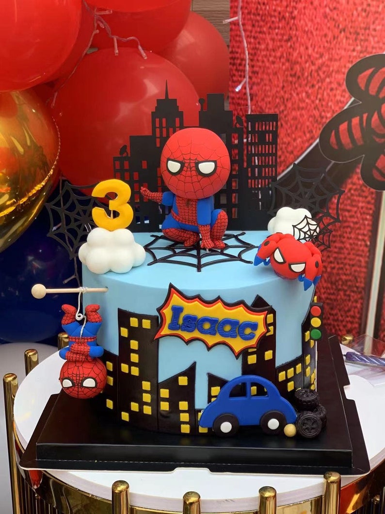 Puede incluir: Un pastel de cumplea&ntilde;os de Spiderman azul y rojo con un fondo de ciudad negra. El pastel est&aacute; decorado con una figura de Spiderman, un Spiderman colgando de una telara&ntilde;a, una ara&ntilde;a roja y negra, un coche azul y el nombre "Isaac" en un bocadillo de estilo c&oacute;mic.