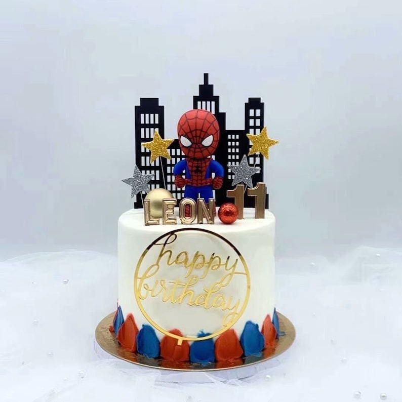 Puede incluir: Un pastel blanco con glaseado rojo y azul. El pastel est&aacute; decorado con una figura de Spiderman, una ciudad, estrellas doradas y las palabras "Feliz cumplea&ntilde;os" y "LEON 11".