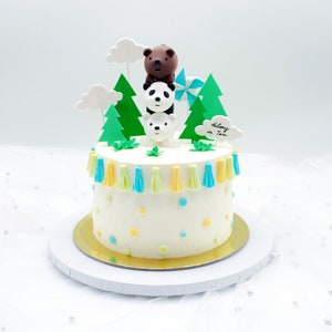 Può includere: Una torta bianca decorata con alberi verdi, nuvole e un orso bruno, un panda e un orso polare. La torta è anche decorata con nappe blu e gialle e puntini colorati. La torta dice "Hilary is Ten" su una nuvola bianca.
