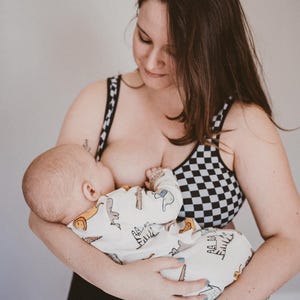 以下が含まれることがあります： 白と黒のチェック柄の授乳用ブラジャーを着た女性が、恐竜柄のロンパースを着た赤ちゃんを抱いています。赤ちゃんは授乳中です。女性は黒いズボンを履いています。