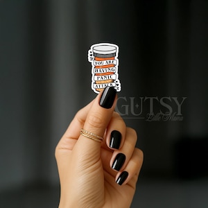 May include: A sticker featuring a pill bottle design with the words "YOU ARE HAVING PANIC ATTACK". The sticker has a white background with orange and black details. The sticker is held by a hand with black nail polish and a gold ring.