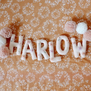 Puede incluir: Letras blancas de fieltro que deletrean el nombre "HARLOW" con pompones rosas y blancos enhebrados en un hilo blanco.