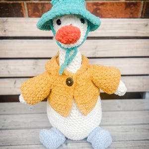 May include: A white crocheted goose plush toy wearing a teal bucket hat with ties, a mustard yellow textured cardigan with a single dark brown button, and light blue crocheted feet. The goose has a bright orange beak and black plastic eyes.
