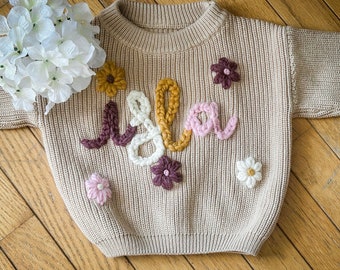 May include: A beige knitted jumper with the name "Isla" embroidered in colourful yarn. The jumper has a crew neck and long sleeves.