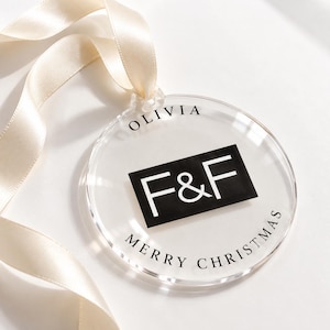 May include: A circular clear acrylic ornament featuring a black rectangular center with the white text F&F. The name OLIVIA is printed in black above the rectangle, and the text MERRY CHRISTMAS is printed in black below. A cream ribbon is attached.