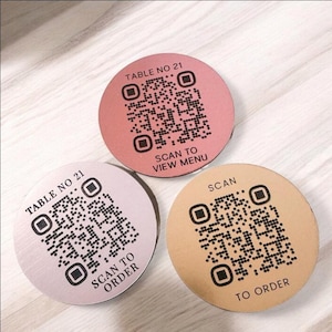 May include: Three circular coasters with QR codes. One is pink with "TABLE NO 21" and "SCAN TO VIEW MENU". Another is light pink with "TABLE NO 21" and "SCAN TO ORDER". The third is tan with "SCAN TO ORDER".