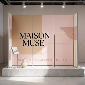 May include: A retail display featuring a large pink and tan backdrop with the black text MAISON MUSE and the phrase MODERN. CONSIDERED. TIMELESS. A gold metal clothing rack stands to the right, with a small sign holder and a cream ceramic vase.