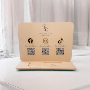 Może przedstawiać: Beżowy stojak wystawowy z logo Premiere Glow Medical Spa i ikonami mediów społecznościowych dla Facebooka, Instagrama i TikToka. Każda ikona ma odpowiadający jej kod QR poniżej. Stojak stoi na białej powierzchni.