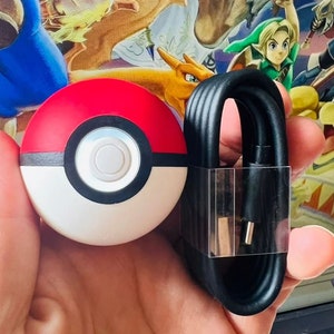 May include: A red and white Poke Ball shaped charging dock for a video game console, with a black charging cable.