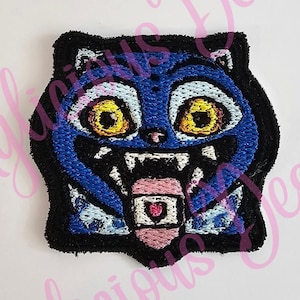 May include: Embroidered patch featuring a cartoon cat with blue fur, yellow eyes, and a pink tongue. The cat has a black outline and is set against a white background. The patch has a playful and slightly menacing expression.