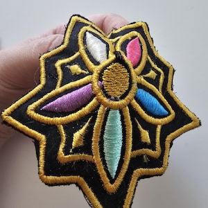 Puede incluir: Parche bordado con un dise&ntilde;o de estrella. El parche presenta un fondo negro con costuras doradas que delinean la forma y los detalles. El centro tiene un c&iacute;rculo dorado con acentos coloridos en rosa, azul, morado, blanco y turquesa.