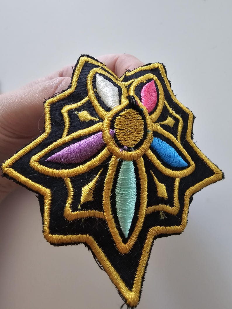 Puede incluir: Parche bordado con un dise&ntilde;o de estrella. El parche presenta un fondo negro con costuras doradas que delinean la forma y los detalles. El centro tiene un c&iacute;rculo dorado con acentos coloridos en rosa, azul, morado, blanco y turquesa.